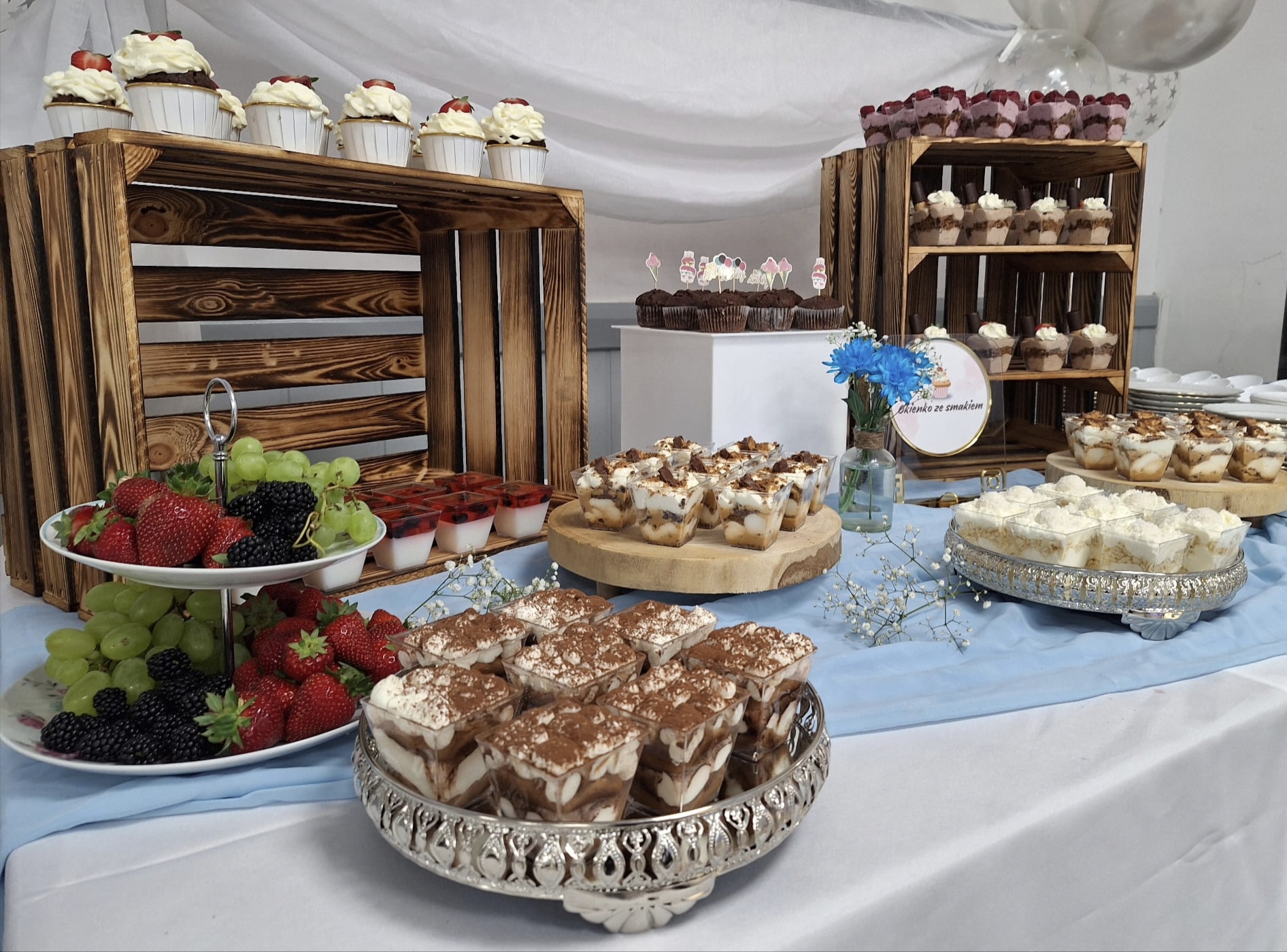 Obficie zastawiony słodki stół w rustykalnym stylu. Desery, w tym tiramisu, panna cotta, babeczki i desery w słoiczkach, ustawione są na paterach oraz na drewnianych skrzynkach. Obok leżą talerze ze świeżymi owocami: truskawkami, winogronami i jeżynami.