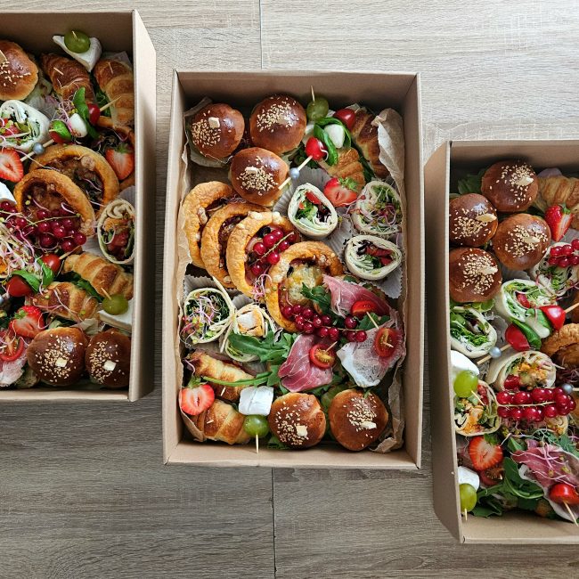 Widok z góry na trzy prostokątne boksy cateringowe wypełnione różnorodnymi przekąskami. W boksach widać mini bułeczki z precla, wrapy, mini croissanty, wytrawne wypieki, koreczki z szynką parmeńską, serem brie, winogronami, truskawkami i porzeczkami.