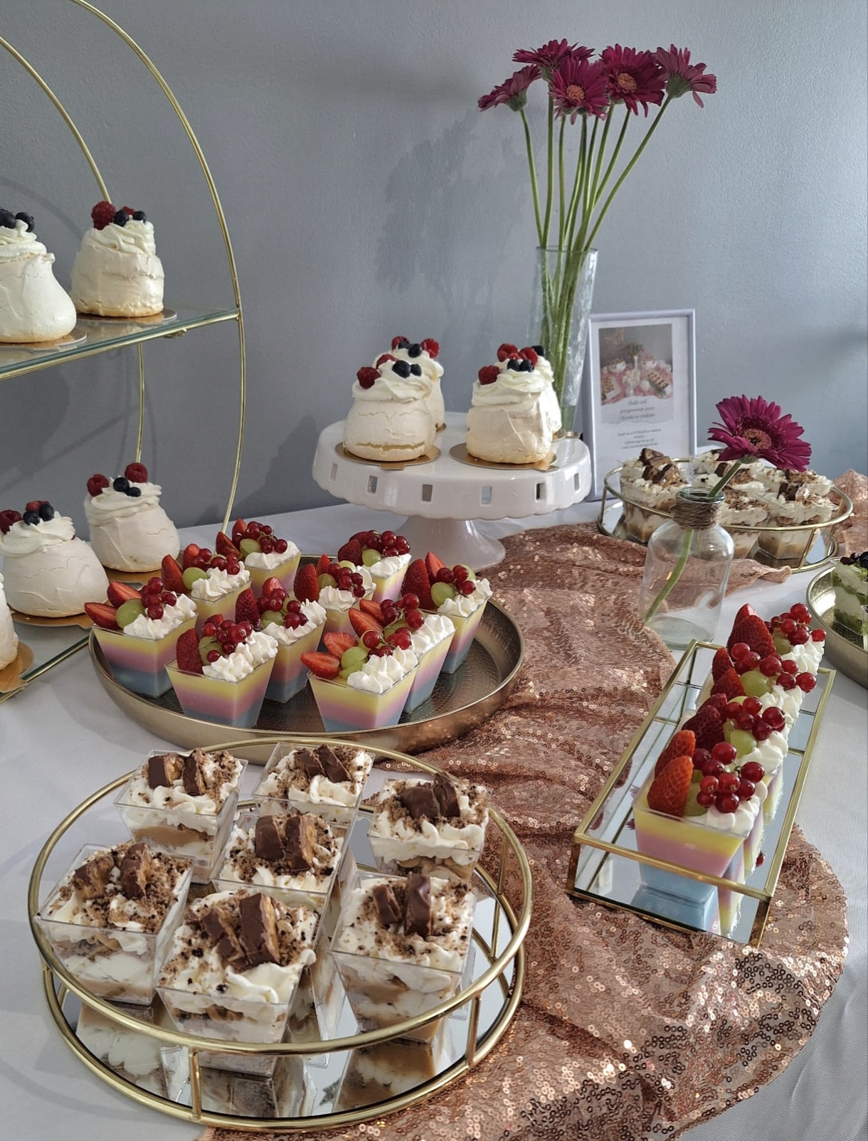 Elegancki słodki stół z deserami. Na złotych paterach i stojakach ustawiono mini bezy pavlova, deserki z kremem i owocami oraz deserki w kubeczkach przypominające tiramisu z kawałkami batonika. Tło stanowi szara ściana i różowe gerbery w wazonie.