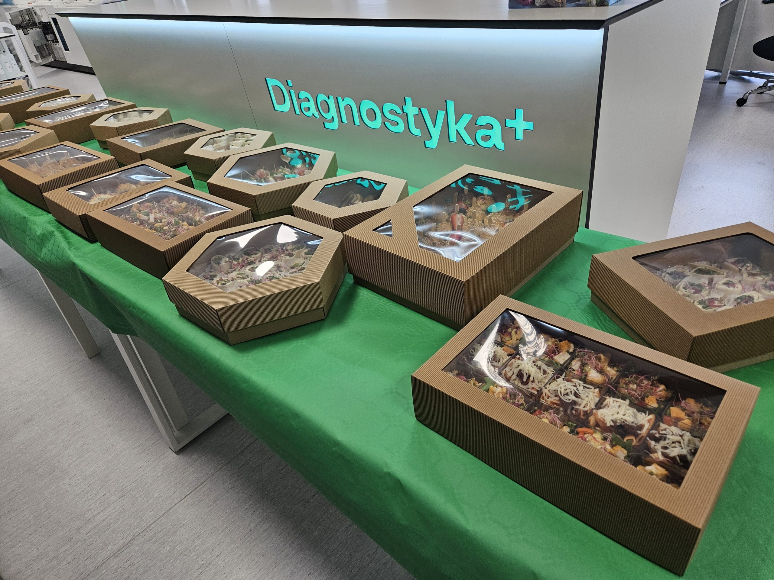 Kilkanaście boksów cateringowych, prostokątnych i sześciokątnych, ułożonych na długim stole przykrytym zielonym obrusem. W tle widoczna jest lada recepcyjna ze świecącym napisem "Diagnostyka+".