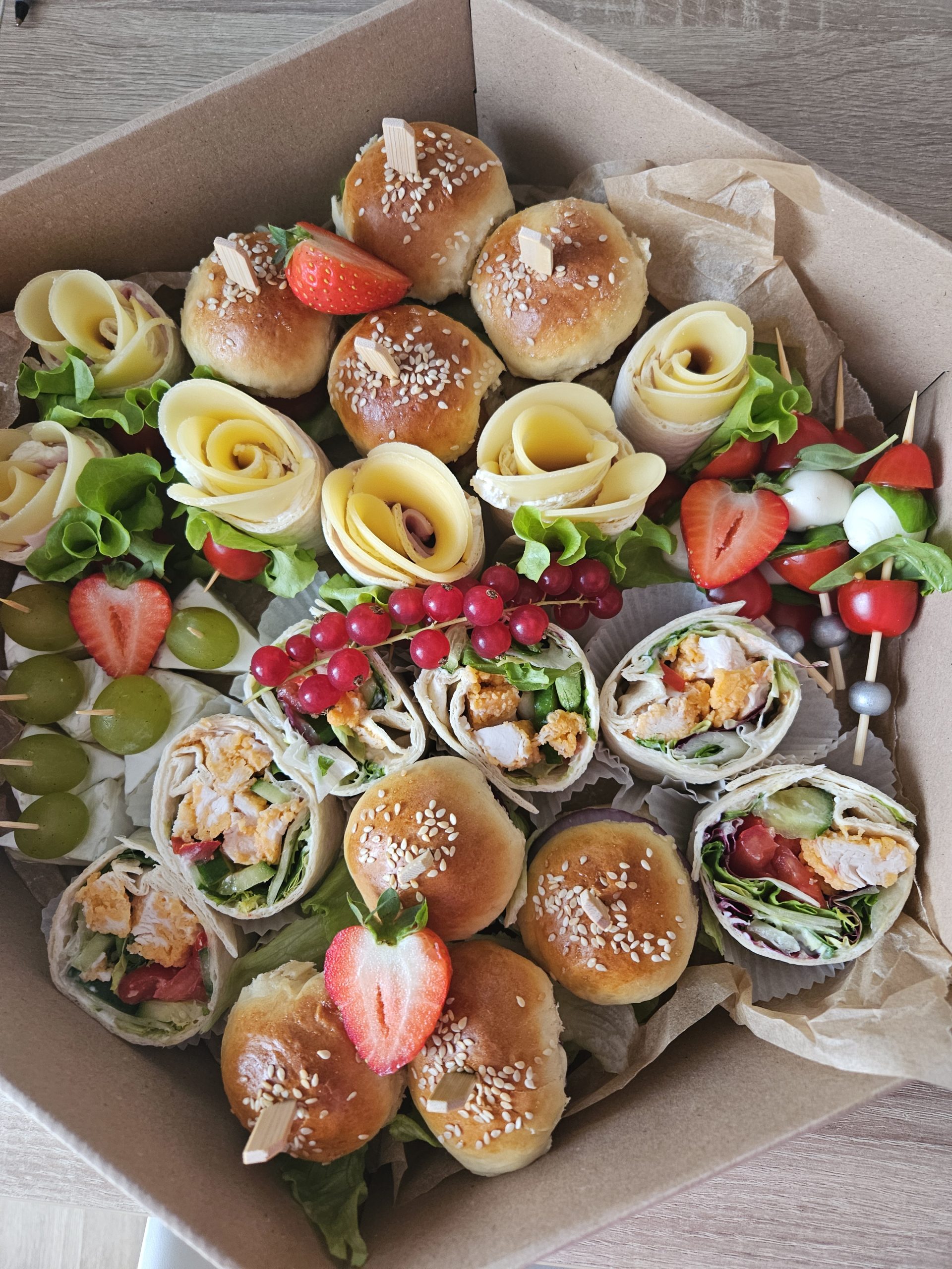 Otwarty boks cateringowy z różnorodnymi przekąskami. Zawiera mini bułeczki z sezamem, roladki z sera w kształcie róży, przekrojone wrapy z kurczakiem i sałatą, szaszłyki caprese oraz owoce, takie jak truskawki, winogrona i porzeczki.