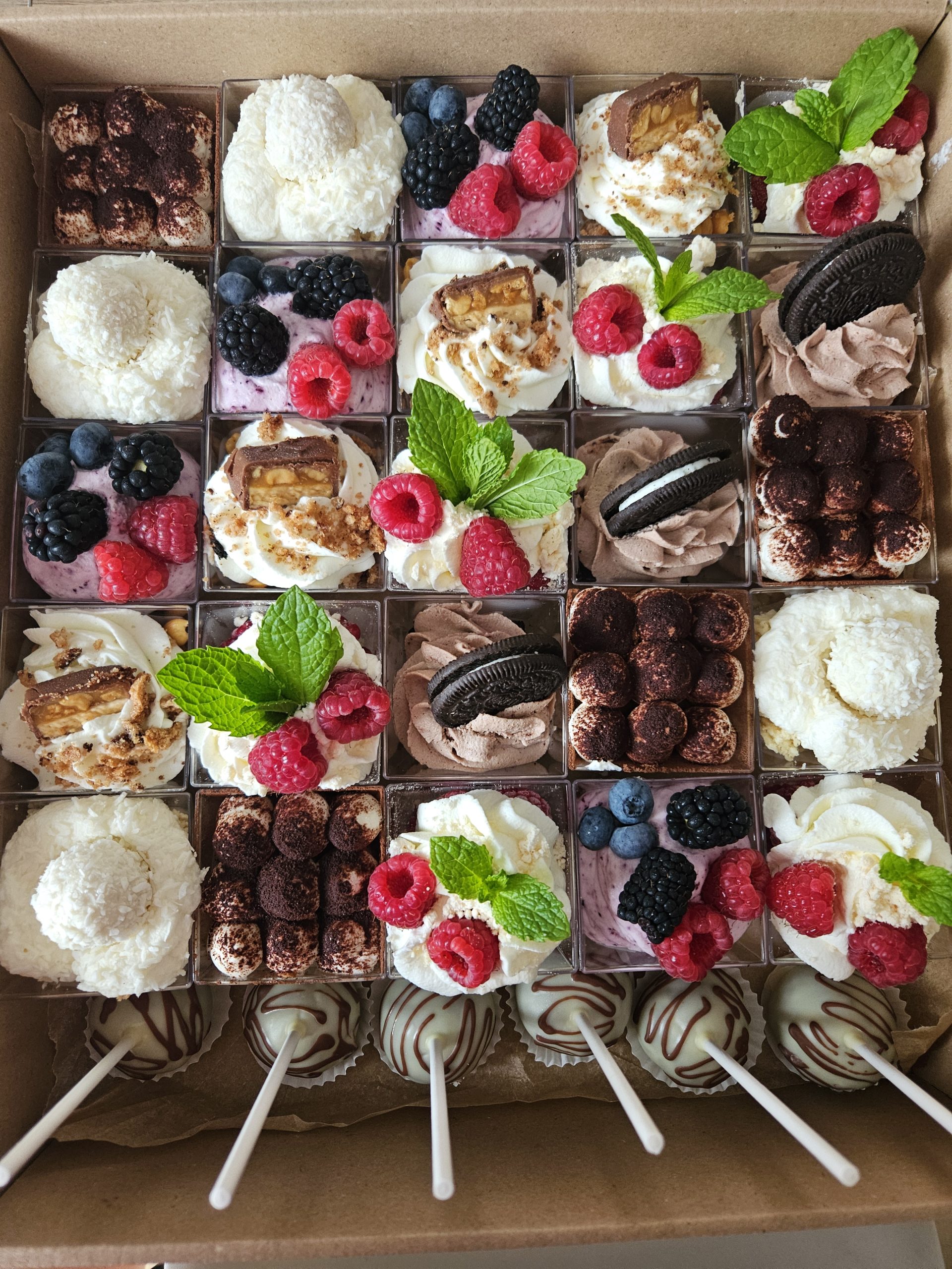 Widok z góry na kartonowy boks wypełniony 25 kwadratowymi mini deserkami. Wśród nich widać tiramisu, deserki kokosowe, deserki z malinami i miętą, deserki typu oreo oraz deserki z kawałkami batonika. Na dole boksu znajduje się 6 cake popsów w białej czekoladzie.