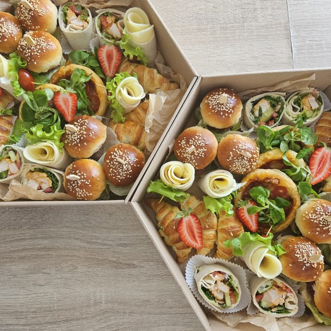 Widok z góry na dwa sześciokątne boksy cateringowe wypełnione przekąskami. W środku znajdują się mini burgery z sezamem, wrapy z kurczakiem, mini croissanty, różyczki zwinięte z sera, sałata oraz połówki truskawek.